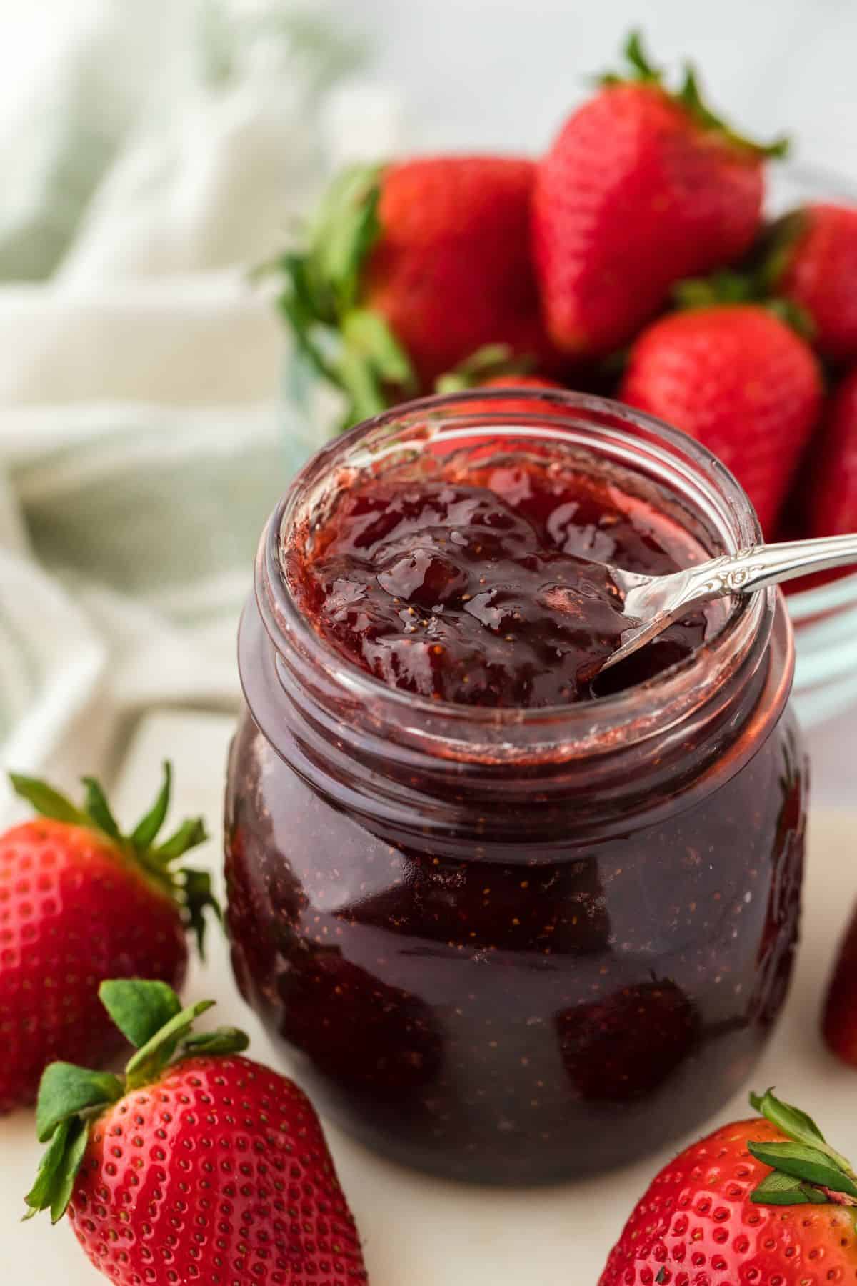 A spoonful of rich strawberry preserves is being lifted from a glass jar, with whole ripe strawberries and a bowl of strawberries in the background