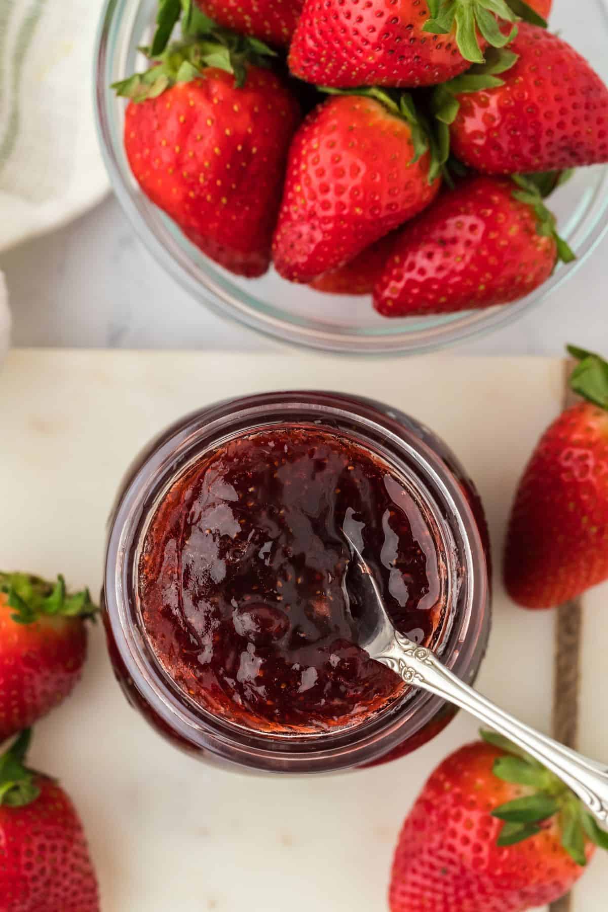 Overhead shot of a glass jar filled with strawberry preserves, with a spoon resting inside it. Surrounding the jar are fresh, whole strawberries in a cutting board and in a glass bowl