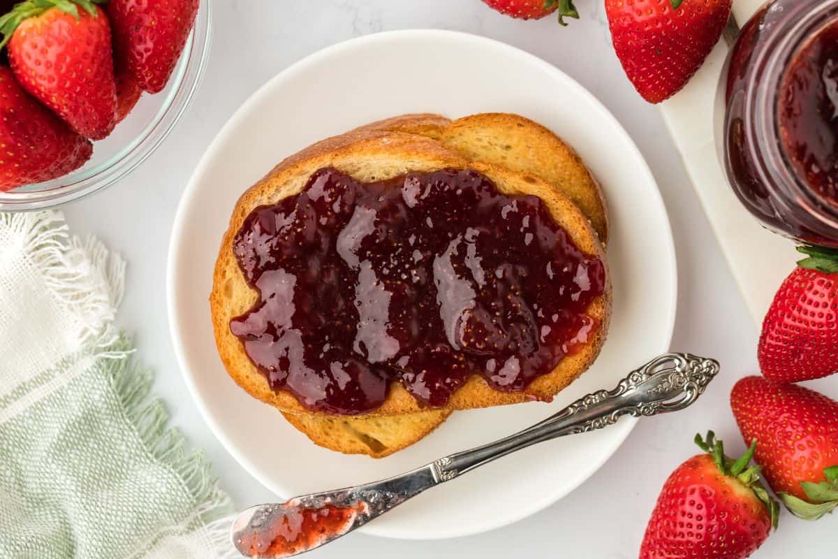 Overhead shot of a piece of toast on a white plate covered with strawberry preserves with a butter knife, accompanied by a jar of preserves and fresh strawberries