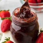A spoonful of rich strawberry preserves is being lifted from a glass jar, with whole ripe strawberries and a bowl of strawberries in the background