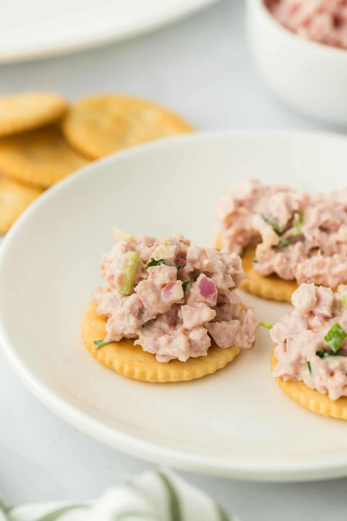 Three round crackers topped with deviled ham, placed on a white plate. Behind them is a bowl with more ham and additional crackers