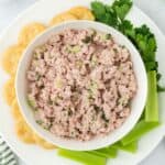 Overhead shot of a bowl of deviled ham surrounded by buttery round crackers and fresh green celery sticks, with parsley garnish to the side