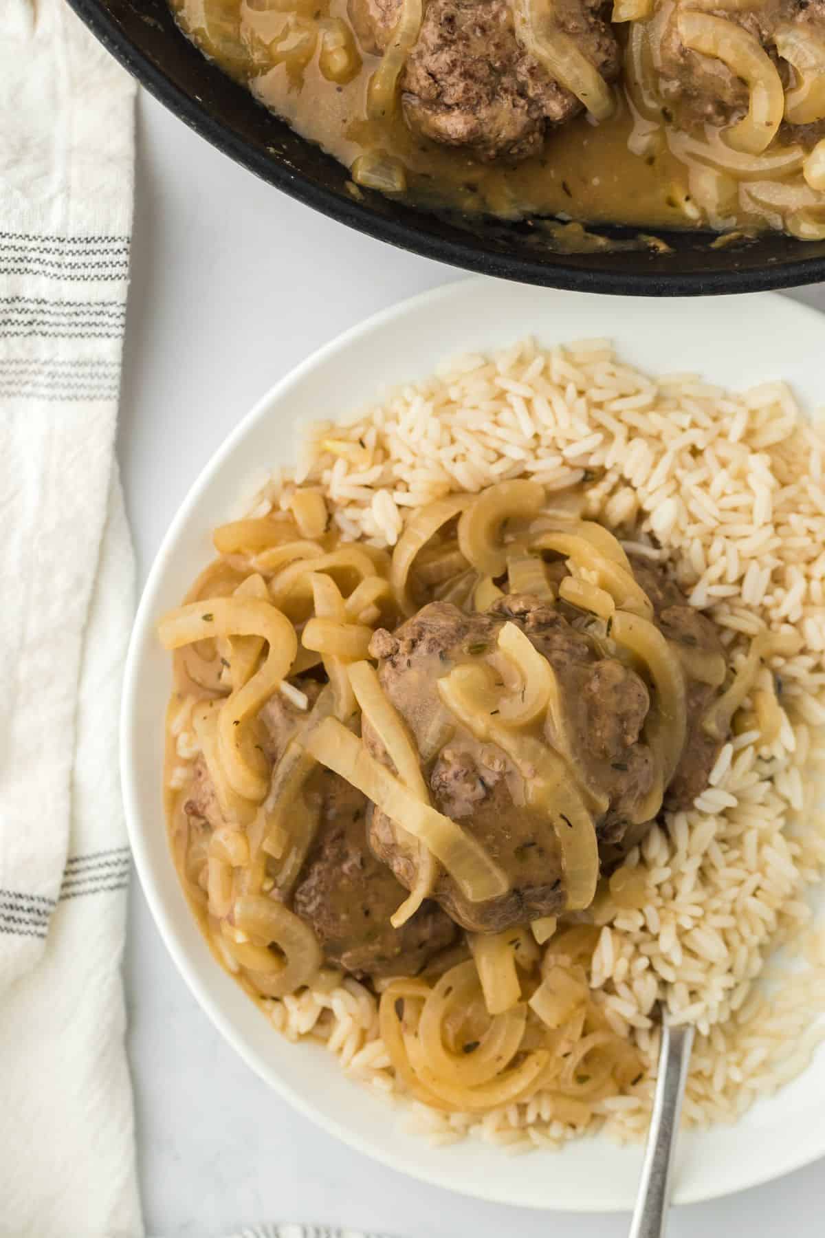 Overhead shot of rice topped with hamburger steaks and sautéed onions, drenched in a rich onion gravy. The dish is accompanied by a fork, and there’s a skillet and a striped towel in the background