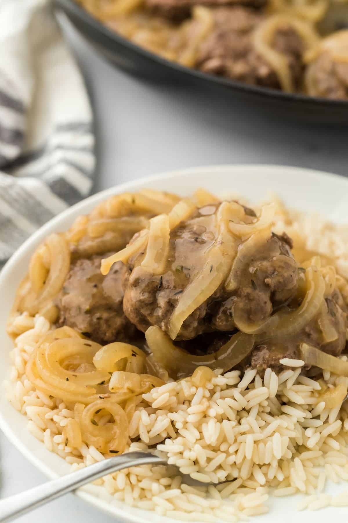 Rice topped with hamburger steaks and sautéed onions, drenched in a rich onion gravy. The dish is accompanied by a fork, and there’s a skillet and a striped towel in the background