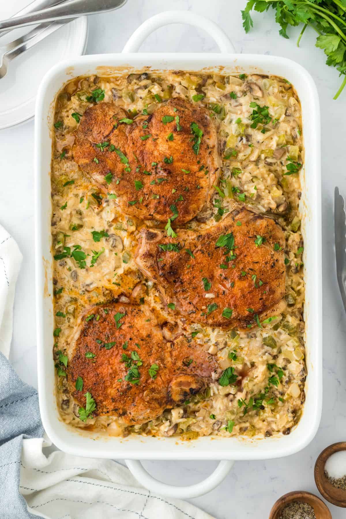 Overhead shot of pork chop casserole in a white rectangular baking dish, garnished with chopped parsley. The dish sits on a marble countertop, accompanied by silver tongs, a cloth napkin, and a fresh bunch of parsley to the side