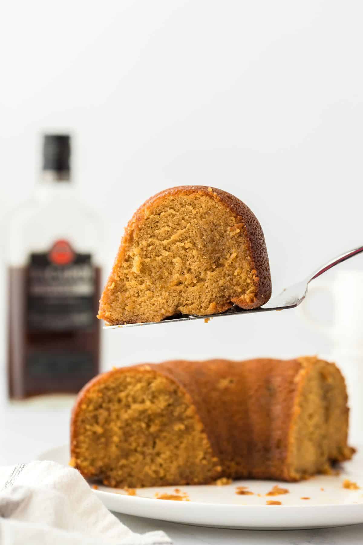A slice of rum cake being held up on a cake server, with the rest of the cake visible on a plate and a bottle of rum in the background