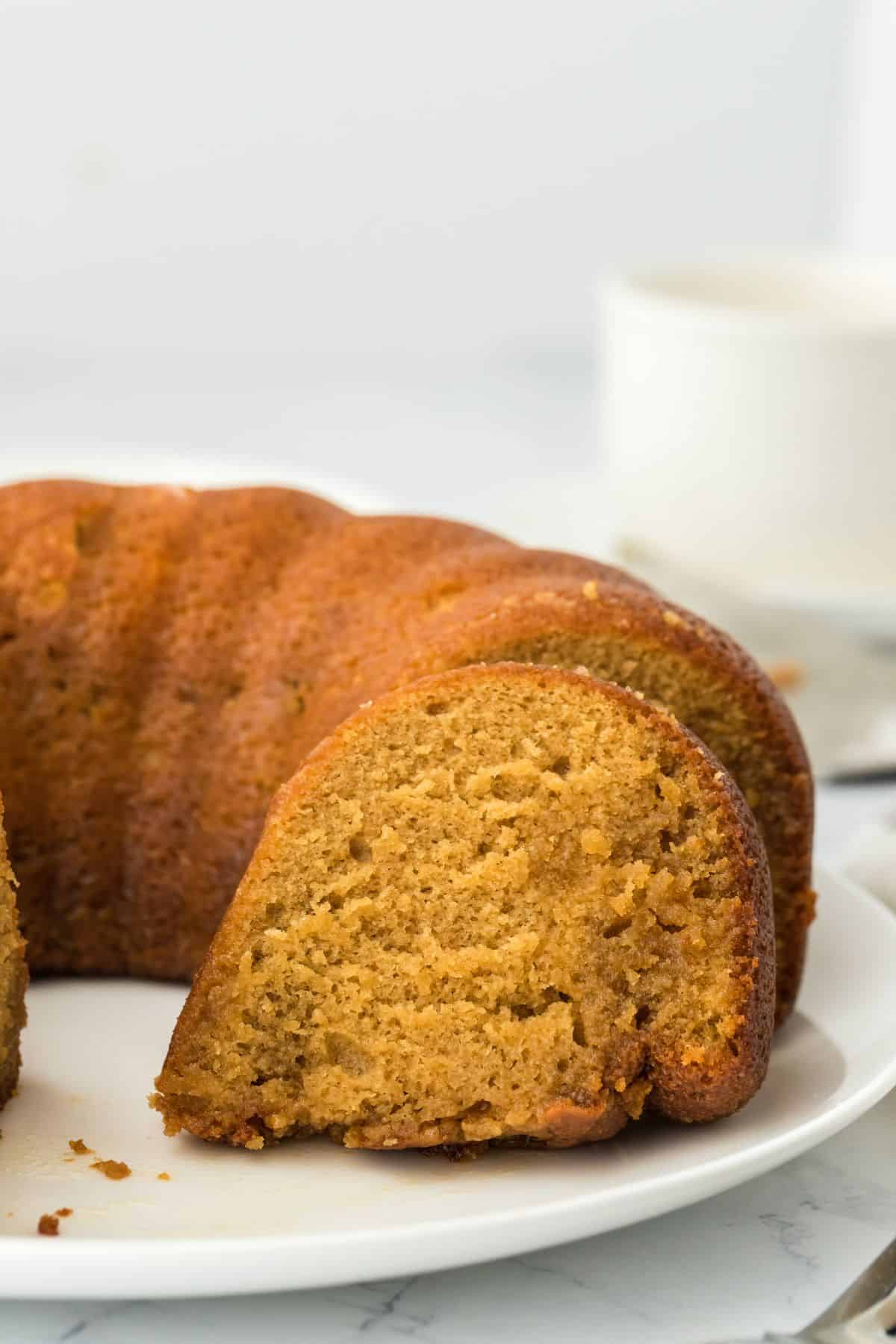 A slice of golden-brown rum cake on a plate, with the rest of the cake in the background, showcasing a moist and airy crumb texture.