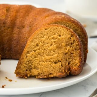 Closeup of a slice of golden-brown rum cake on a white plate, with the rest of the cake in the background