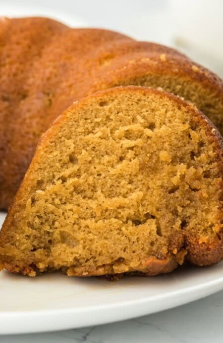 Closeup of a slice of golden-brown rum cake on a white plate, with the rest of the cake in the background