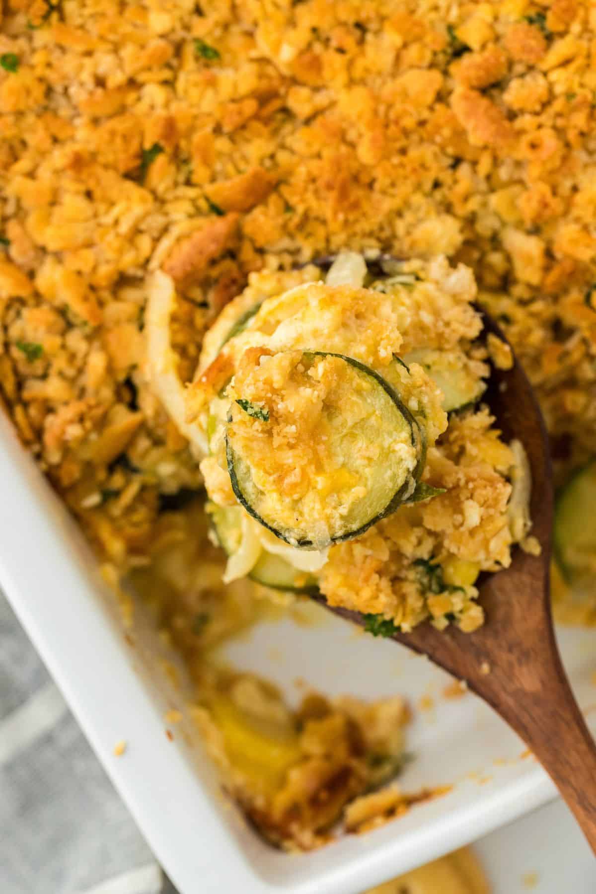 Close up of a baked squash casserole in a white dish, topped with a crispy, golden-brown crust. A wooden spoon is scooping a portion, revealing the creamy interior filled with slices of yellow squash and zucchini