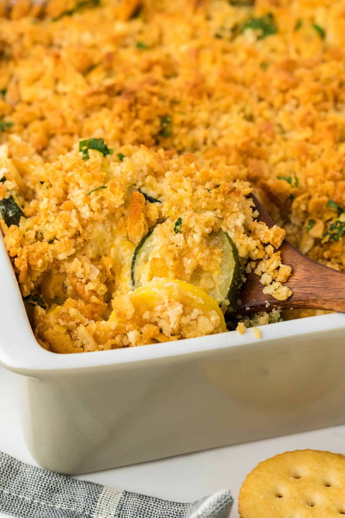 Close up of a baked squash casserole in a white dish, topped with a crispy, golden-brown crust. A wooden spoon is scooping a portion, revealing the creamy interior filled with slices of yellow squash