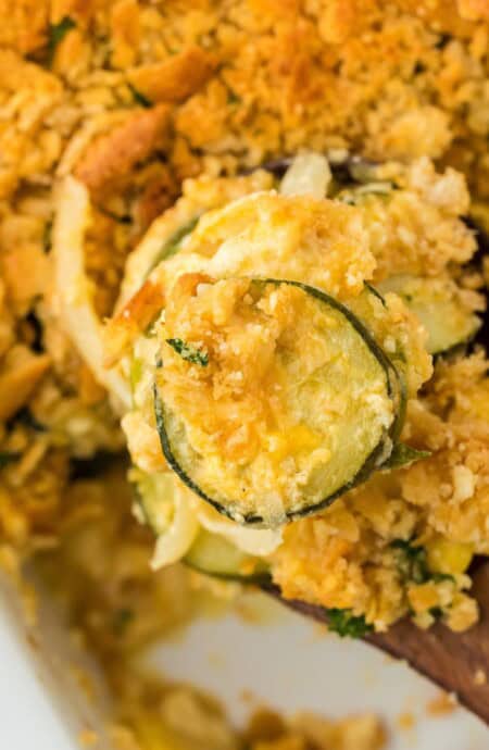 Close up of a baked squash casserole in a white dish, topped with a crispy, golden-brown crust. A wooden spoon is scooping a portion, revealing the creamy interior filled with slices of yellow squash and zucchini