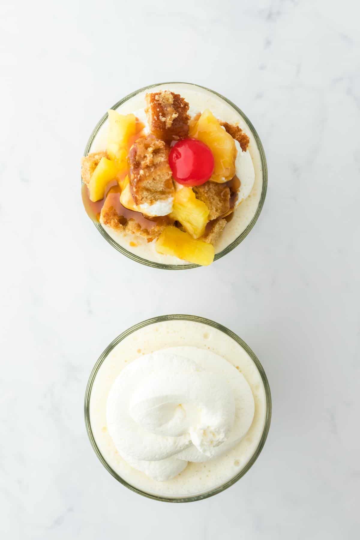 Two pineapple milkshakes topped with whipped cream. One garnished with pineapple chunks, crumbled cake, caramel drizzle, and a maraschino cherry