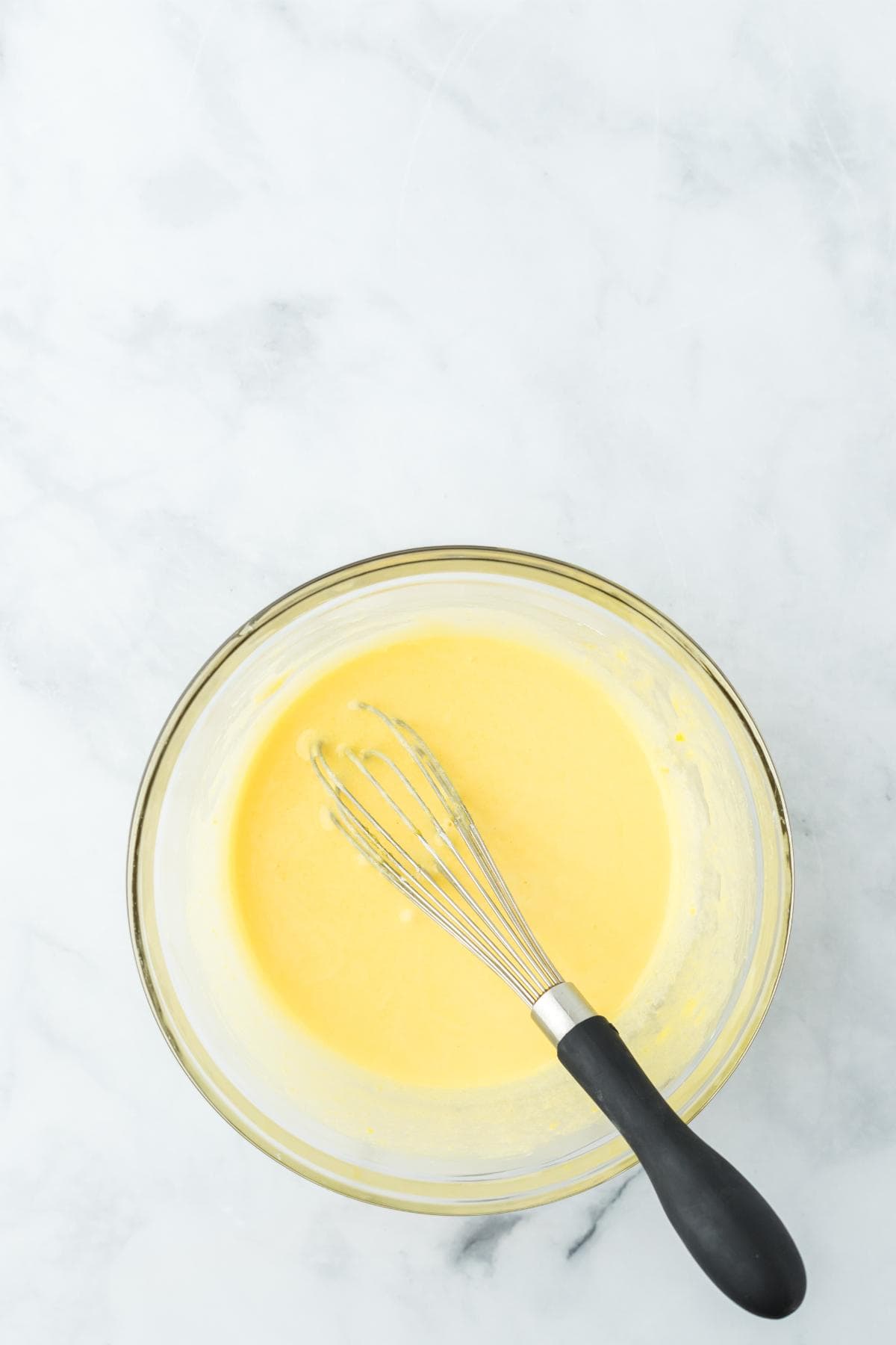 A clear glass bowl contains a mixture of egg yolks, sugar, cornstarch, and salt