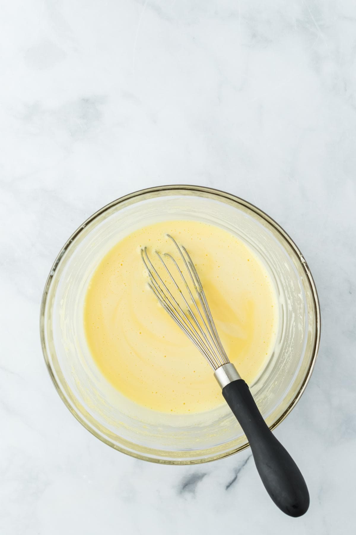 A clear glass bowl with the tempered custard mixture, with a whisk resting inside