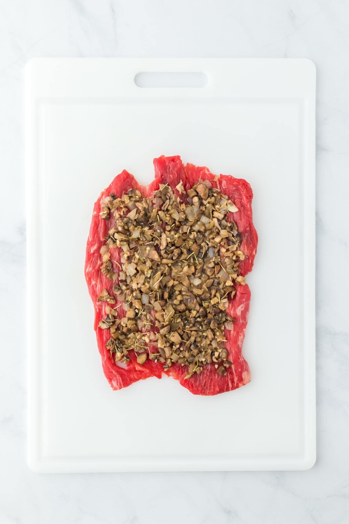 Raw flank steak laid flat on a cutting board, topped with the cooked mushroom mixture