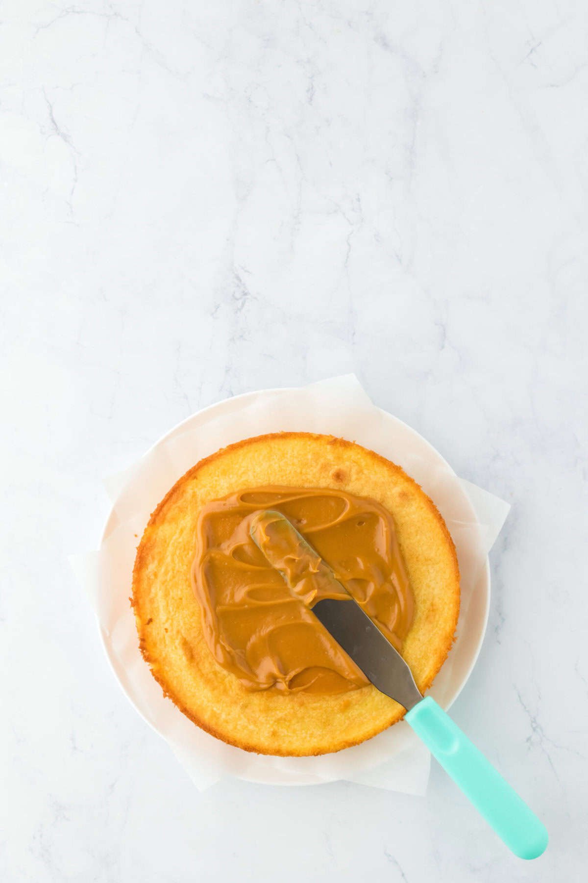 Layer of yellow cake being spread with caramel icing with spatula on white countertop