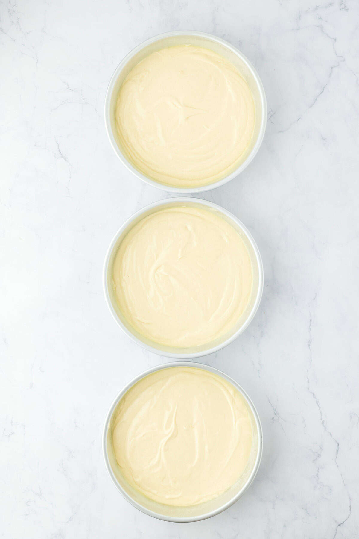 Three cake pans filled with yellow cake before baking on white countertop