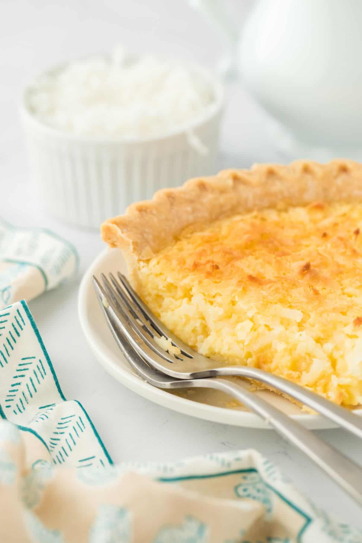 Close up of a slice of coconut custard pie on a plate with a fork. In the background there is a bowl of shredded coconut