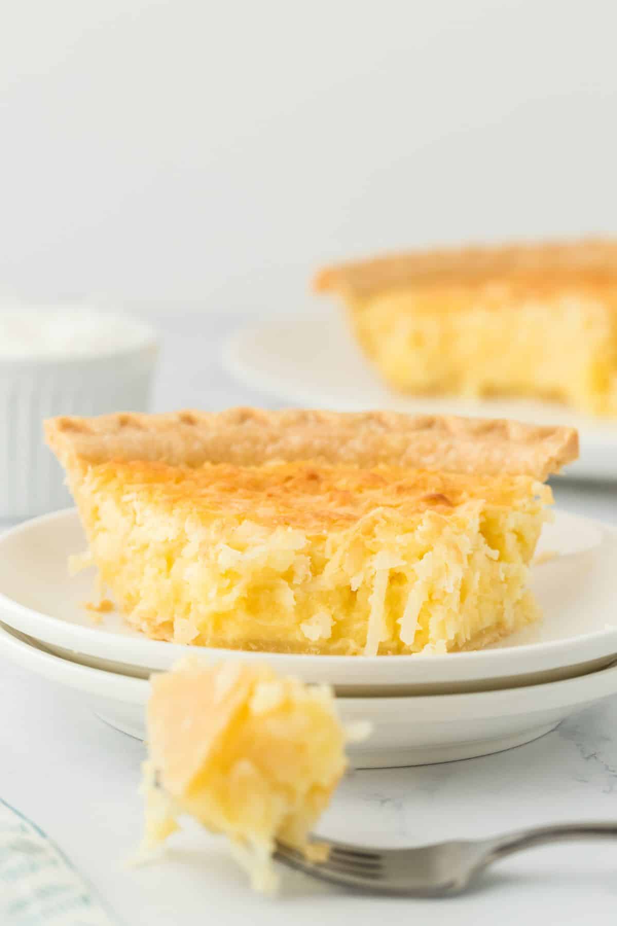 A slice of coconut custard pie on a white plate with a fork in front of it piercing a piece. Another slice is in the background