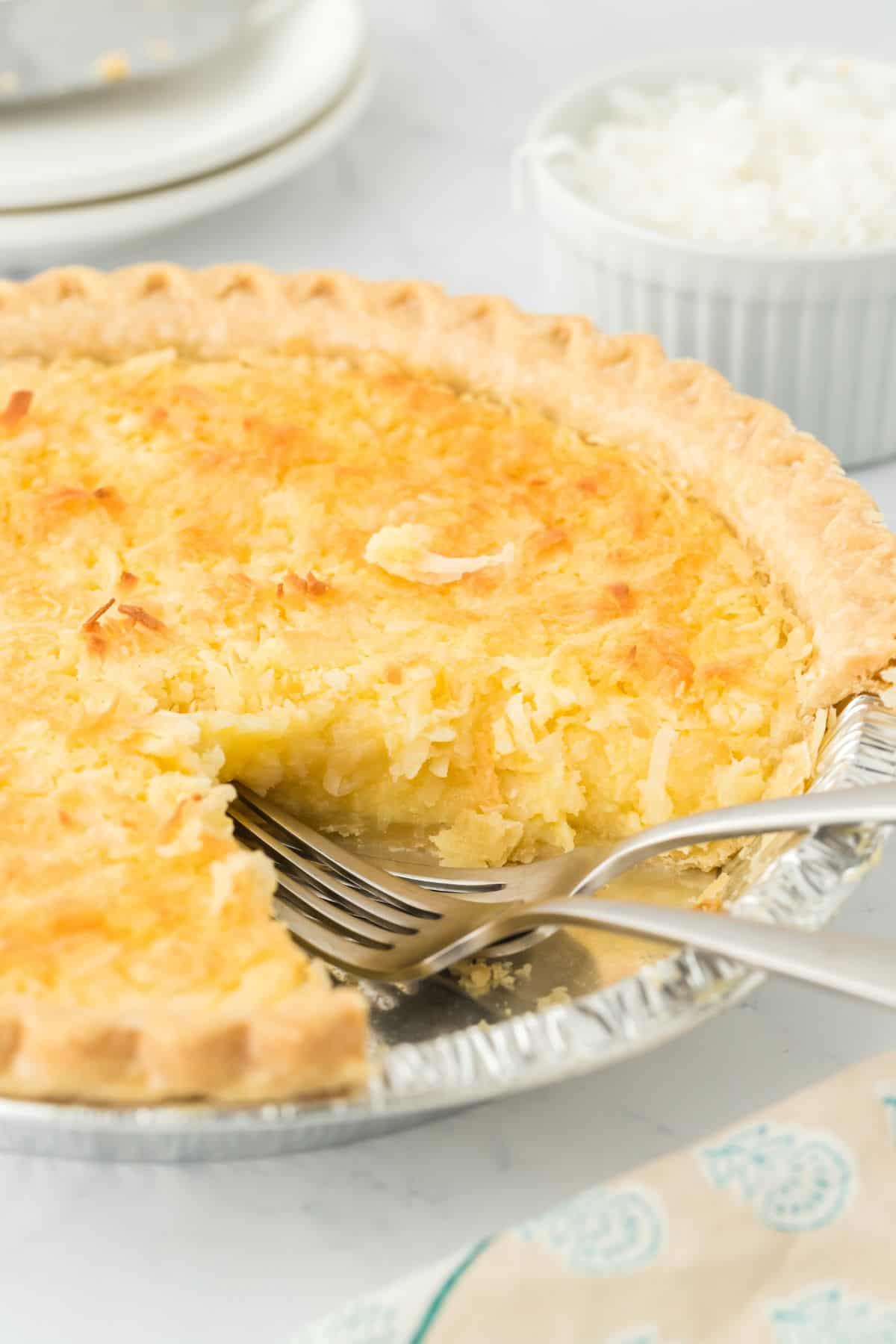 Closeup of a coconut custard pie with a slice removed, revealing its creamy interior. A fork is in the pie tin, and a bowl of shredded coconut is in the background