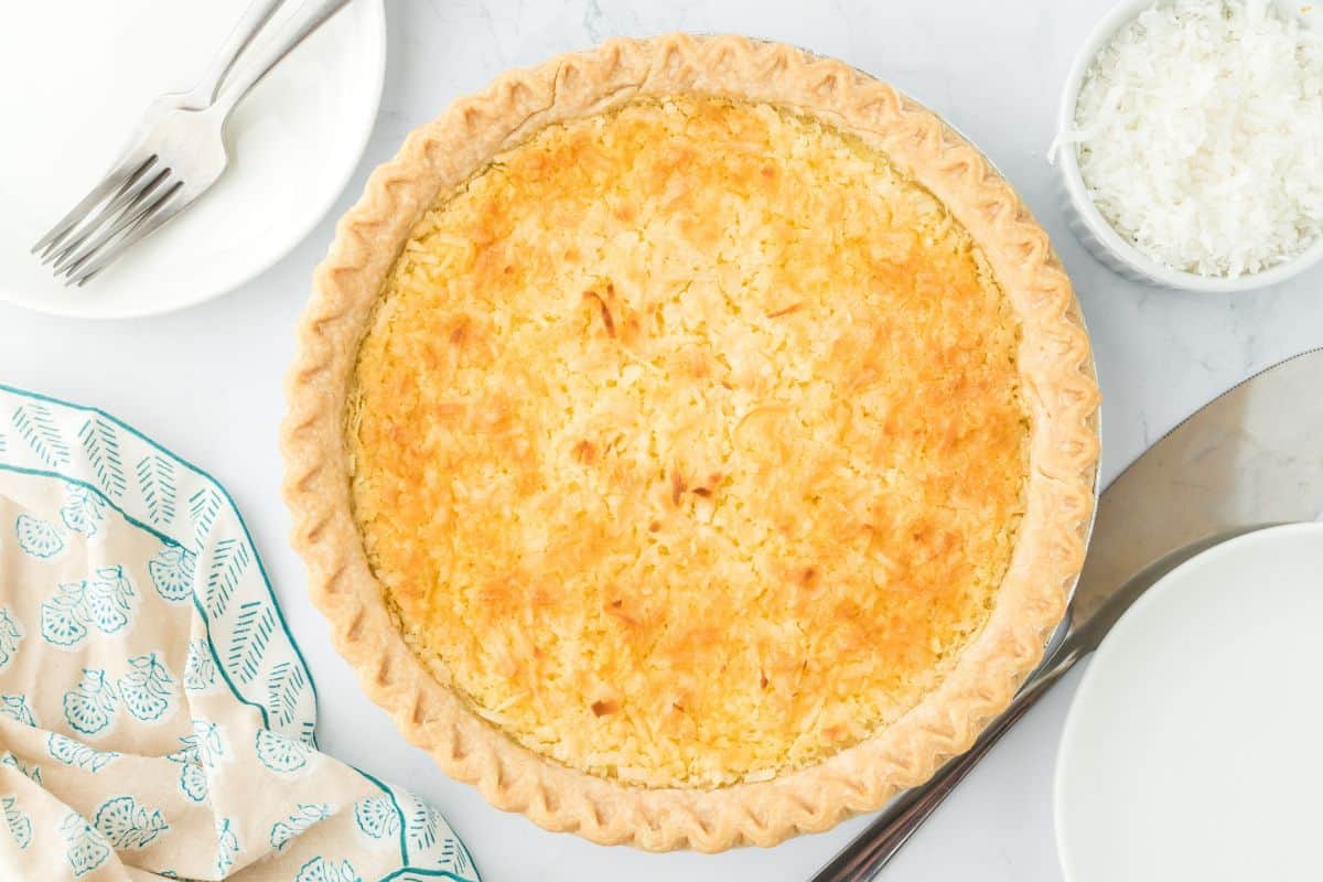 Overhead shot of a whole coconut custard pie with a golden crust and a bowl of shredded coconut nearby. Plates, a kitchen cloth and forks are set beside the pie