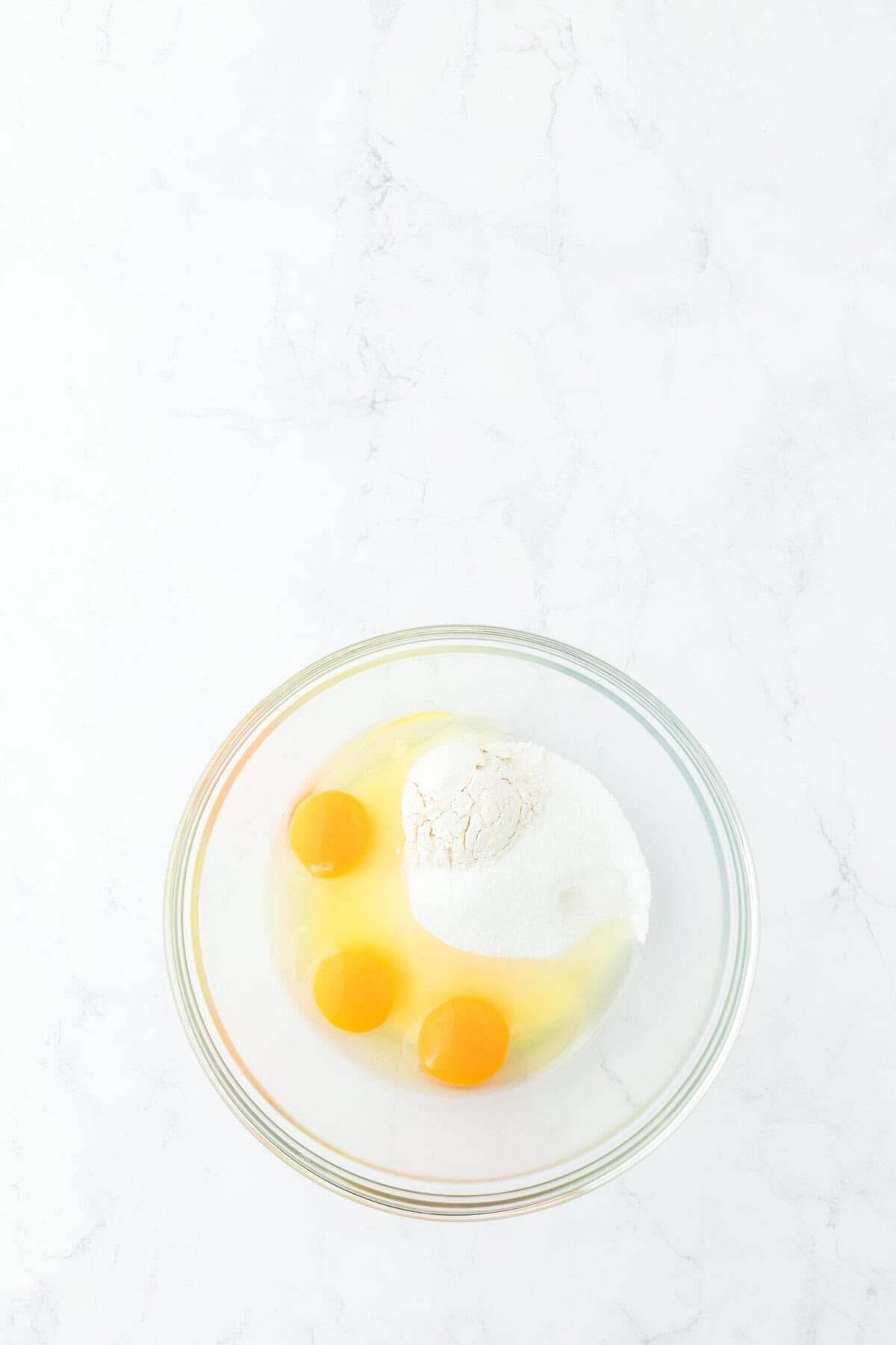 Eggs, granulated sugar, flour and extract in a glass bowl on white countertop