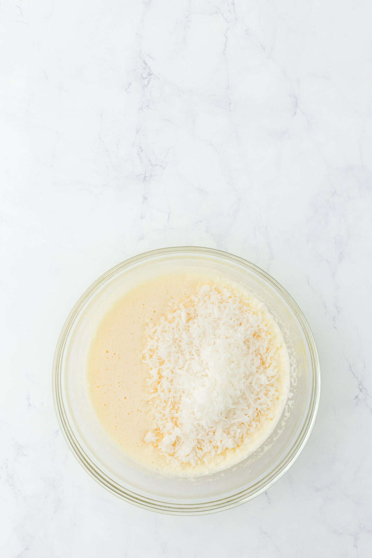 Coconut flakes added to a glass bowl of custard pie filling on white countertop.