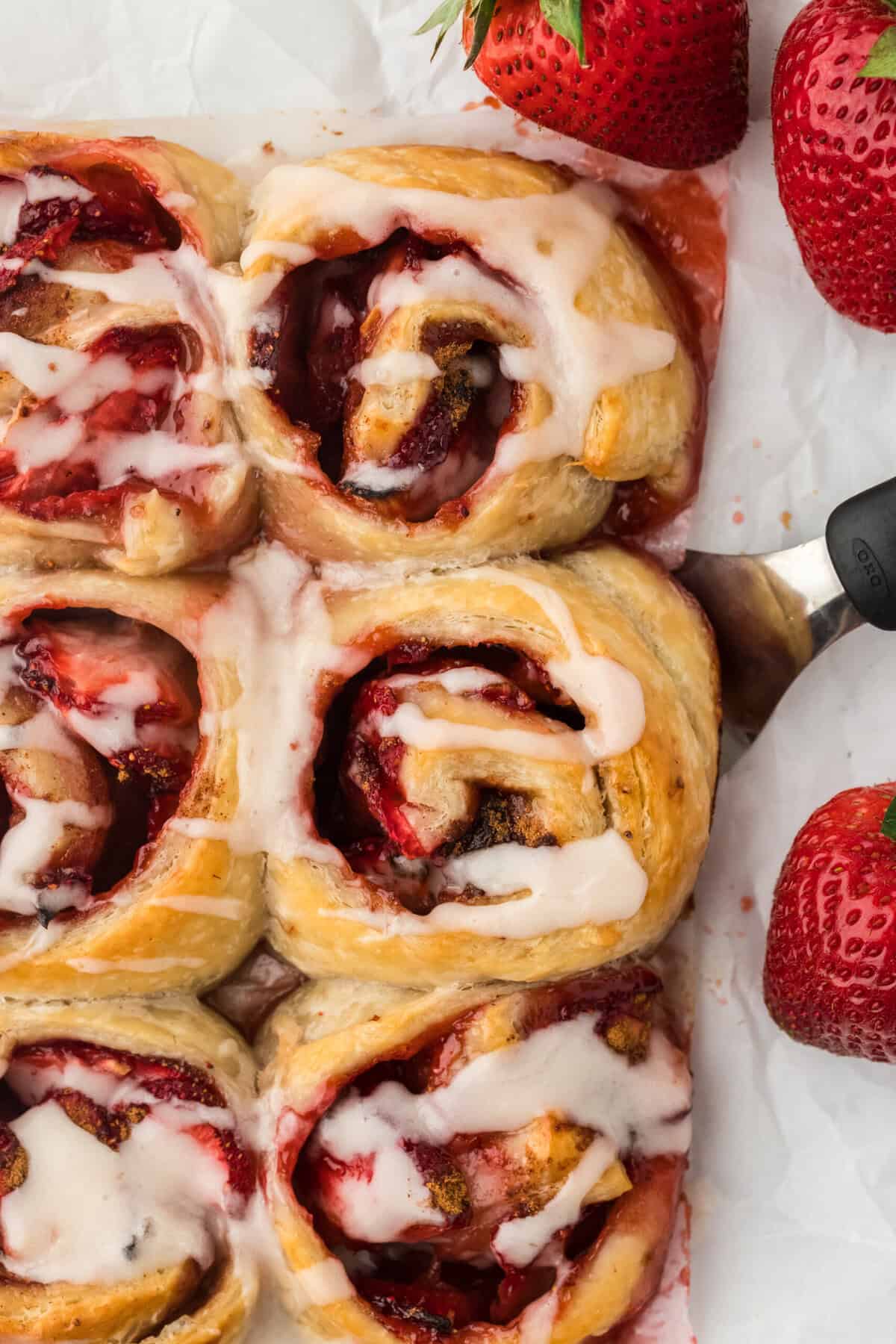 An overhead of strawberry cinnamon roll recipe with a spatula removing one with strawberries in the background