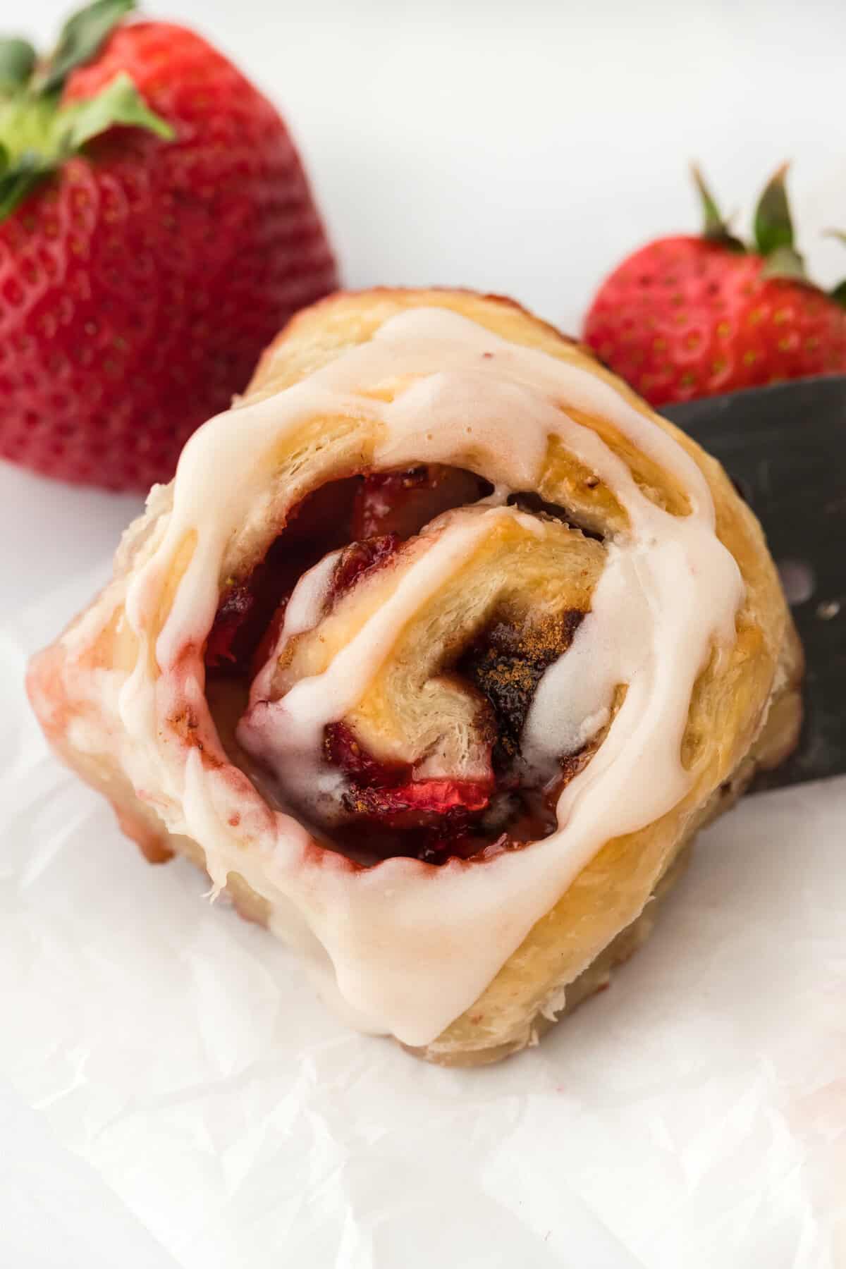 A close up of a puffy pastry strawberry cinnamon roll with two strawberries in the white backgound