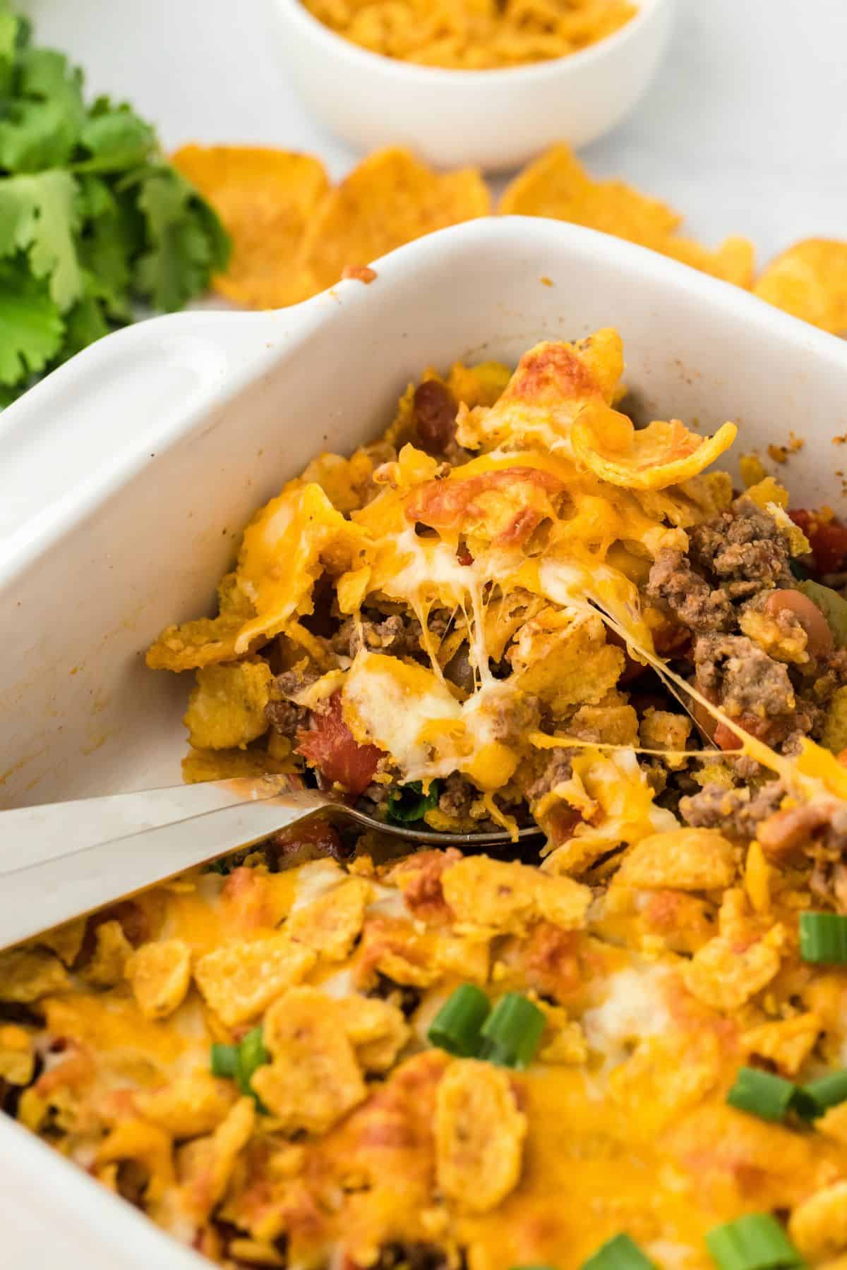 A close-up of a serving of Frito pie being scooped from a casserole dish, showing layers of ground beef, melted cheese, Fritos corn chips, and tomatoes. Fresh cilantro and a bowl of Fritos are in the background
