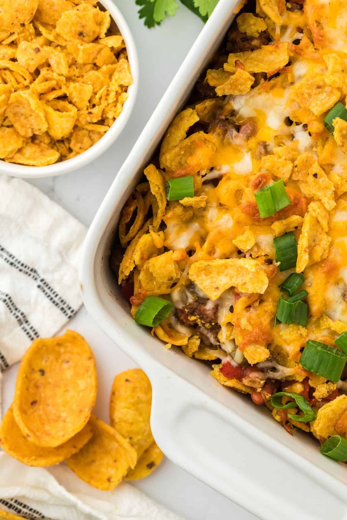 Overhead shot of a baked Frito pie with ground beef, melted cheese, Fritos corn chips, and chopped green onions. A bowl of Fritos and some loose chips are next to the dish