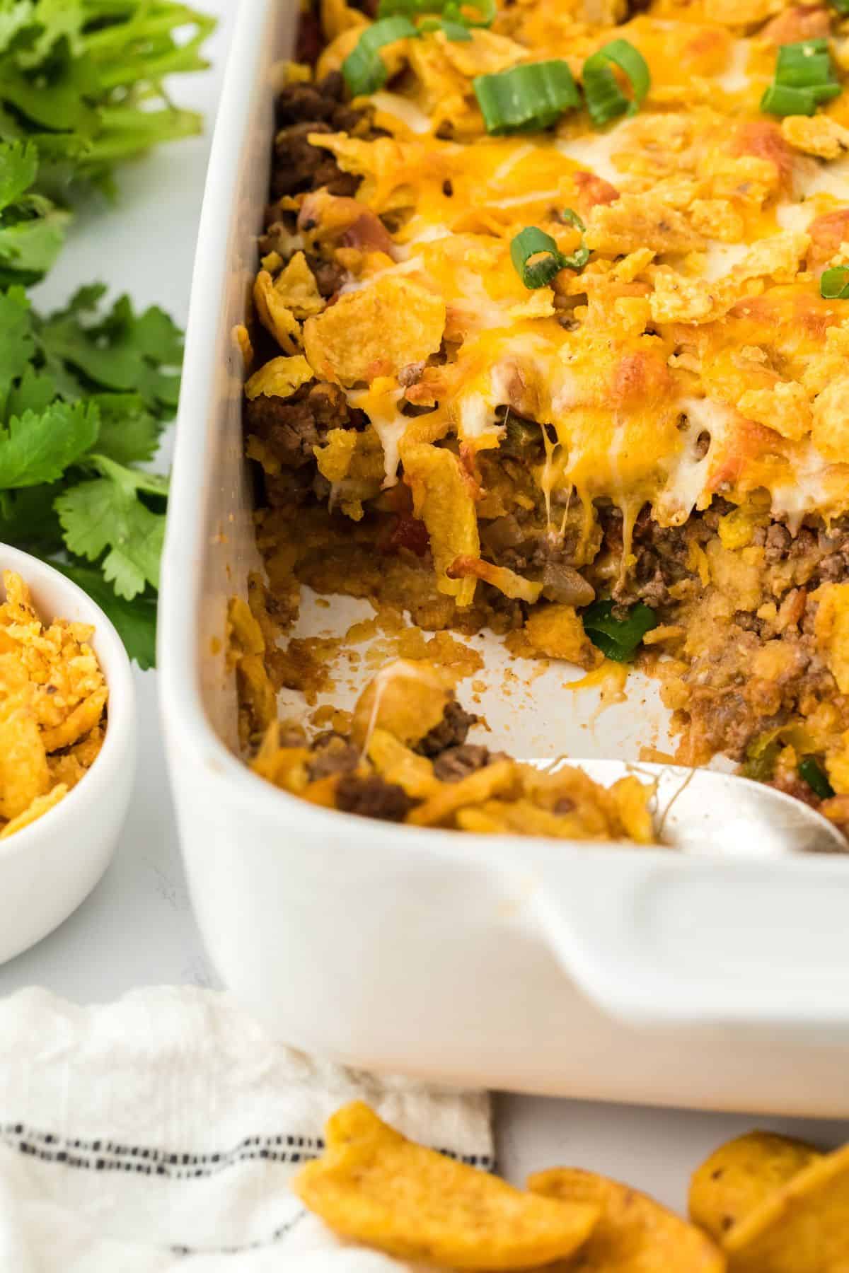 Close-up of a partially eaten Frito pie in a casserole dish, showing layers of ground beef, melted cheese, Fritos corn chips, and chopped green onions. Fresh cilantro and a bowl of Fritos are in the background