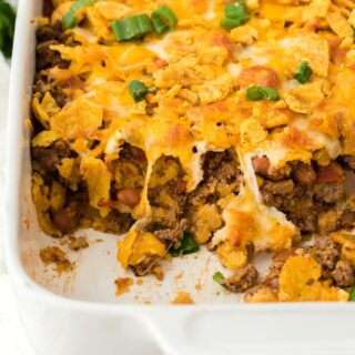 Close up of a partially eaten Frito pie in a white baking dish, showing layers of ground beef, melted cheese, Fritos corn chips, beans, and chopped green onions, with a striped napkin next to it