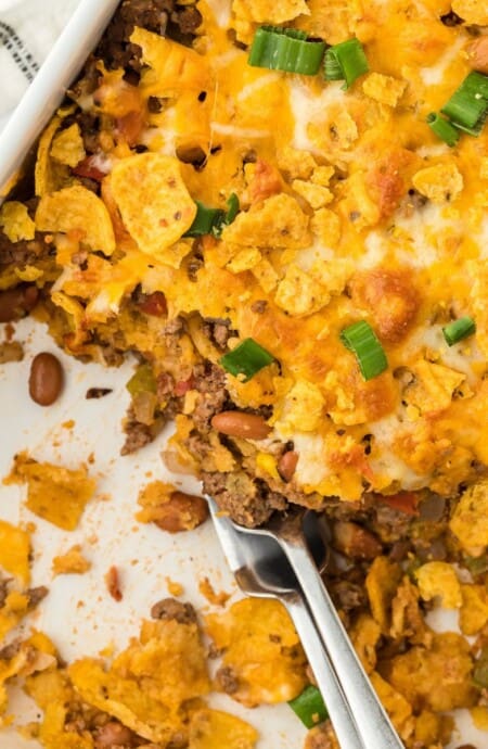 Overhead shot of a partially eaten Frito pie in a casserole dish, showing layers of ground beef, melted cheese, Fritos corn chips, beans, and chopped green onions. A serving spoon is in the dish, and a white and black striped napkin is nearby