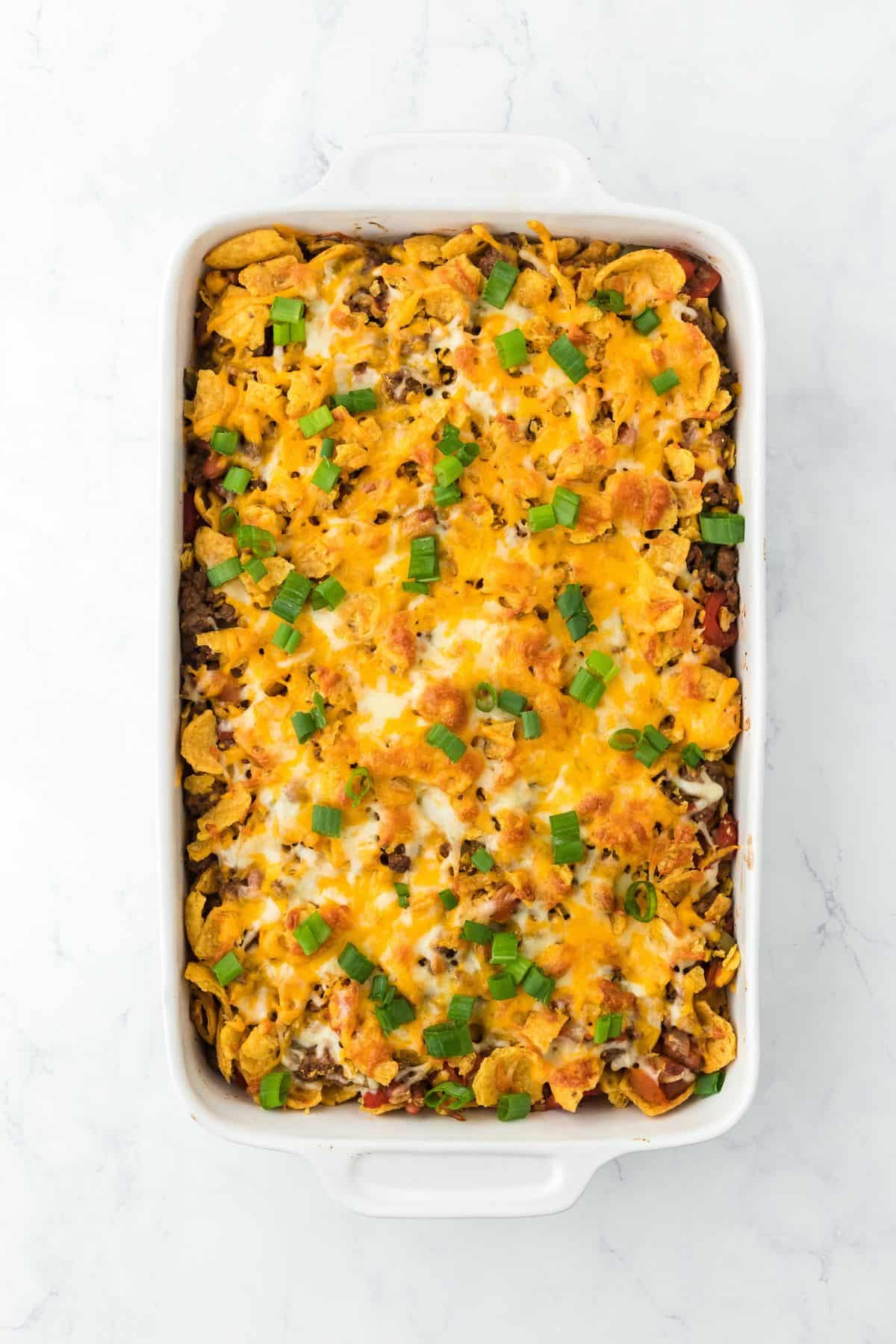 Overhead shot of a baked Frito pie in a casserole dish, topped with melted cheese, Fritos corn chips, and chopped green onions. A bowl of Fritos, a fork, a spoon, and fresh cilantro are placed nearby
