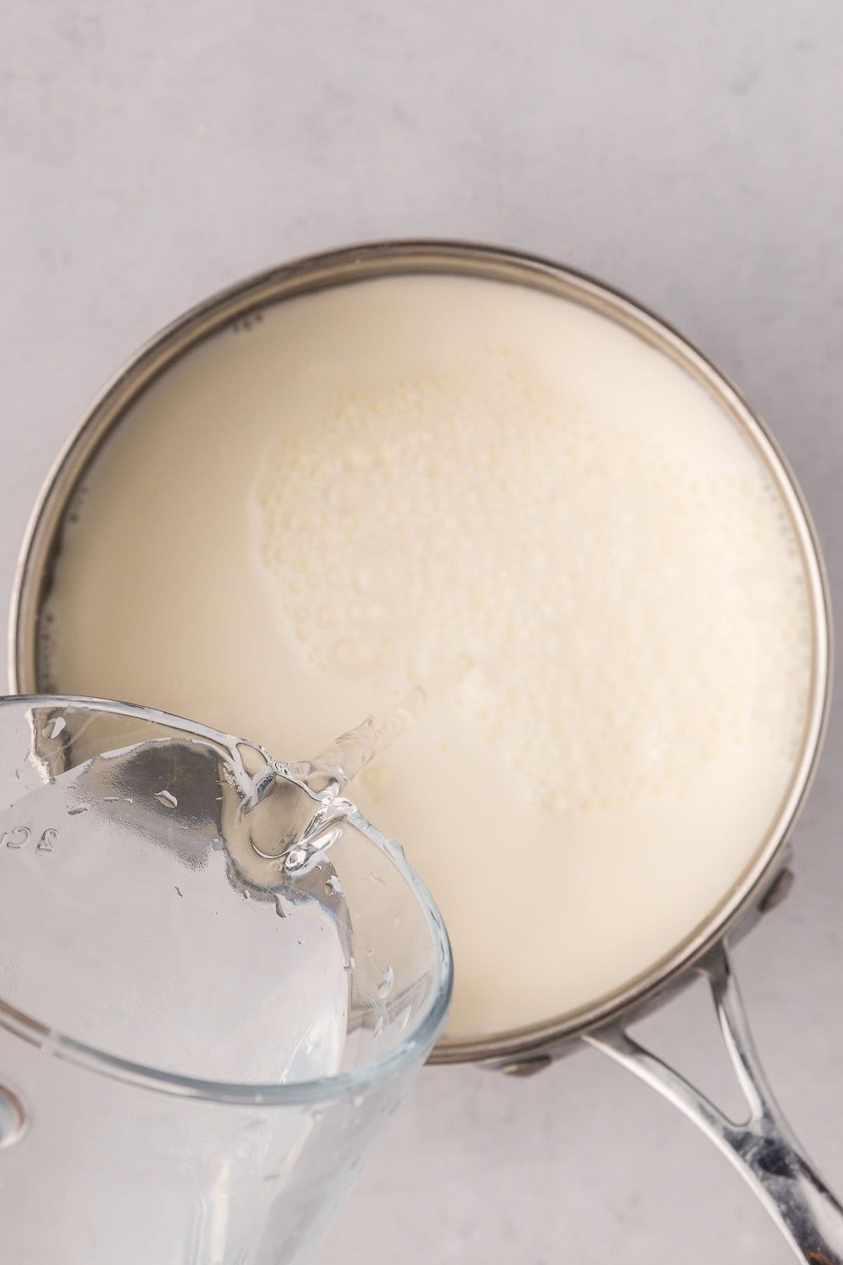 A glass measuring cup pouring water into a pot of half and half on the stove, ready to start grits