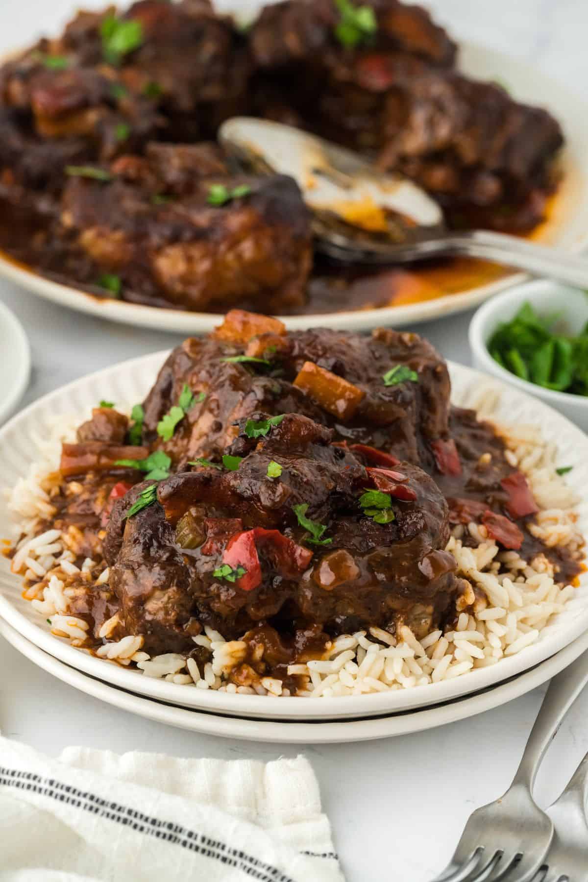 A plate of Jamaican oxtail stew over rice, with more oxtail pieces on a serving platter in the background and a bowl of parsley nearby