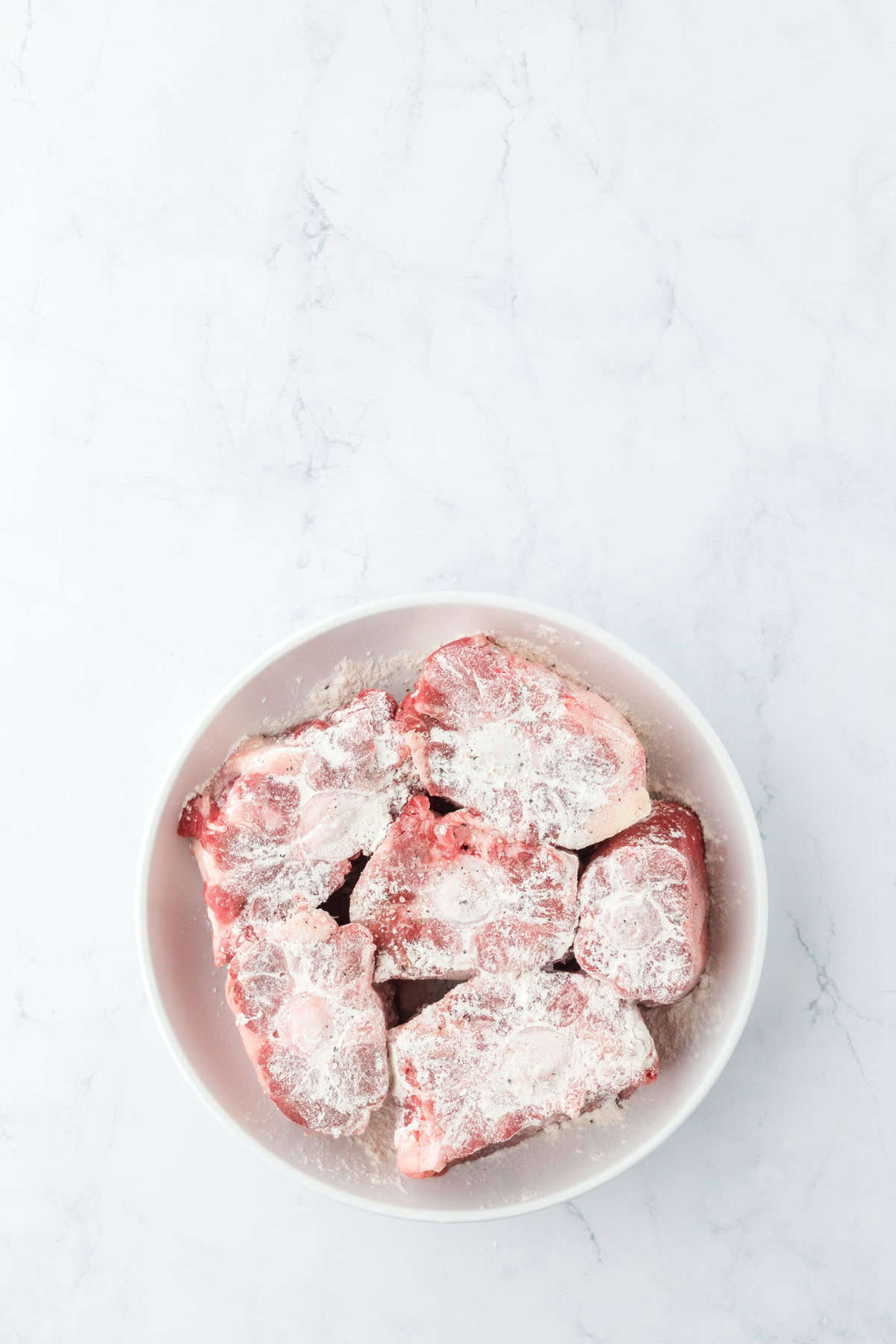 Seasoned and lightly floured oxtails on a white plate on white countertop