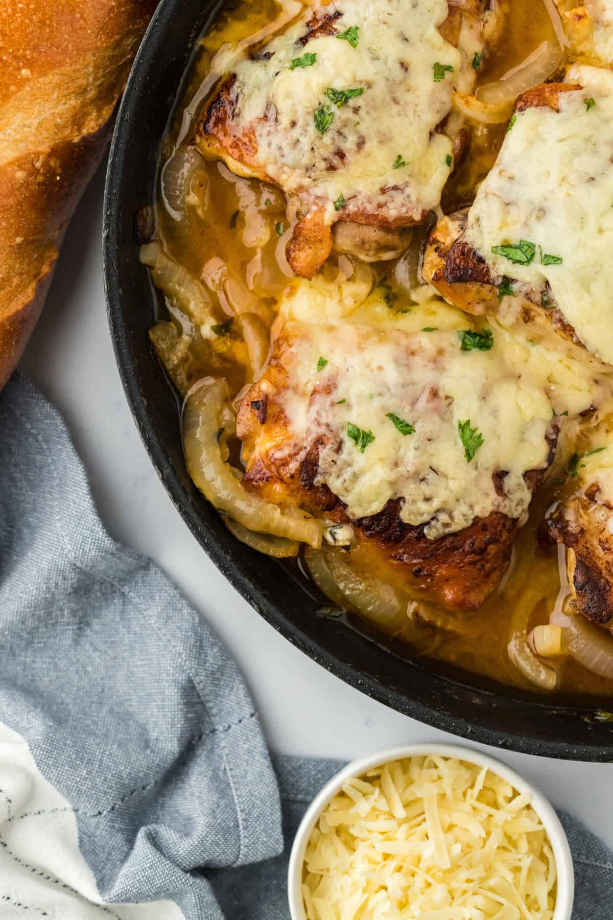 Closeup of french onion chicken in a skillet on a white background. Next to it there are ingredients like shredded cheese, and toasted baguette slices