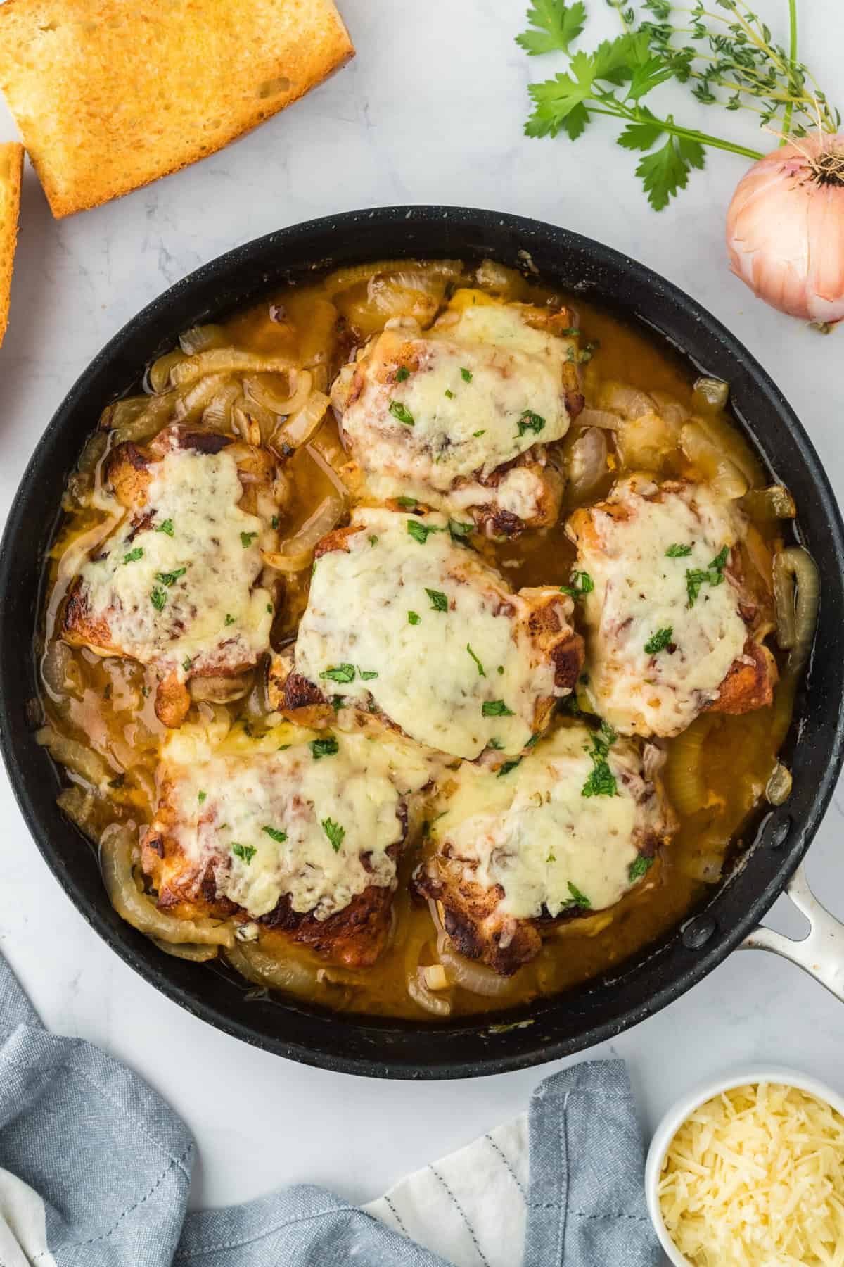 Overhead shot of french onion chicken in a skillet on a white background. In the background, there are ingredients like onions, garlic, fresh parsley, a bunch of fresh thyme, shredded cheese, and toasted baguette slices