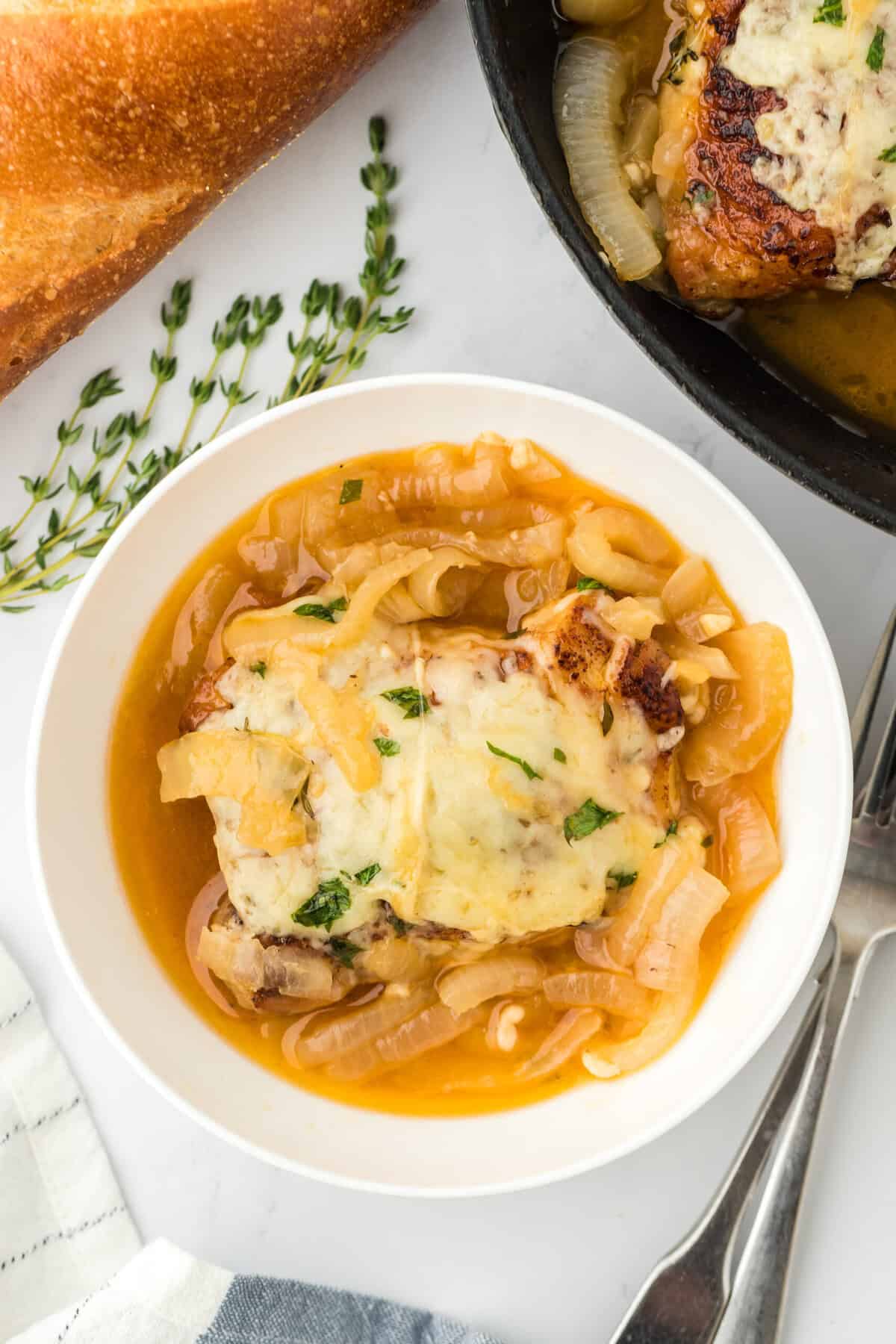 French onion chicken in a white bowl, topped with melted cheese and a generous amount of caramelized onion broth. In the background, there's a baguette, fresh parsley, and a fork