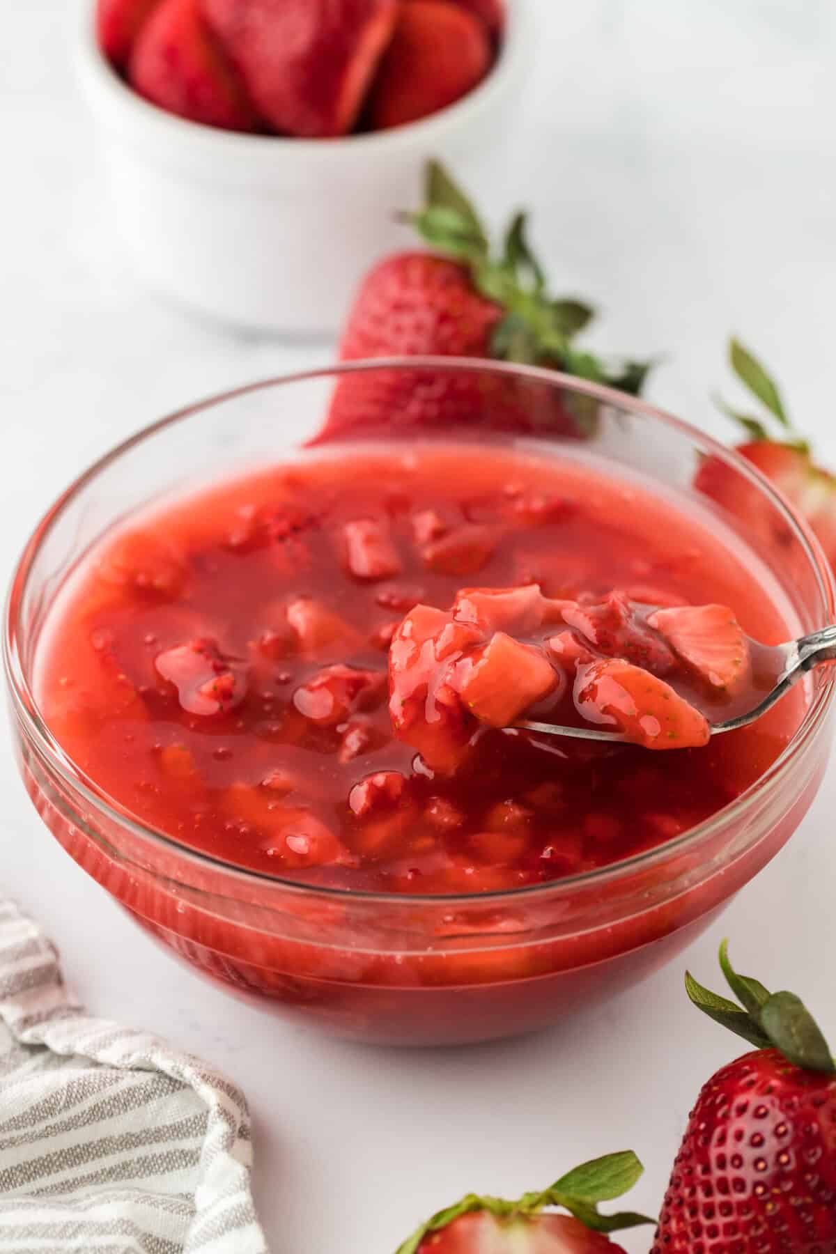 A spoon lifting a portion of strawberry sauce from a clear glass bowl. The bowl is surrounded by whole and halved fresh strawberries.