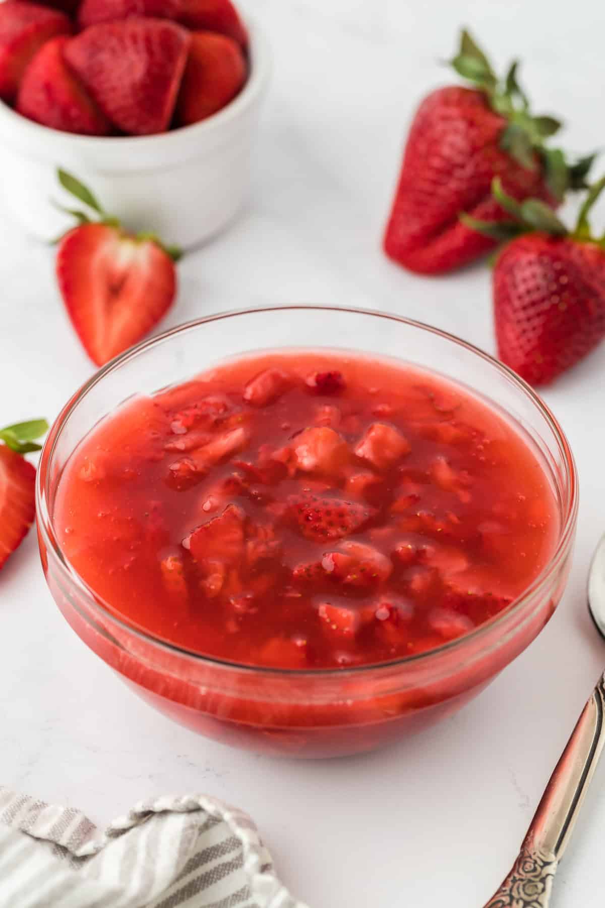 Strawberry sauce in a clear glass bowl with a spoon by it. The bowl is surrounded by whole and halved fresh strawberries on a wooden surface. In the background, there's a white white bowl with more strawberries