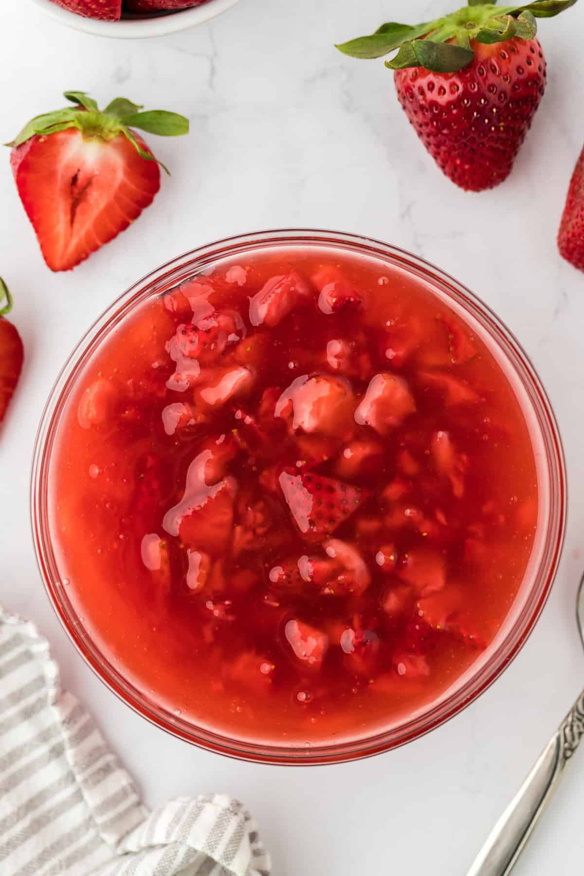 Closeup of a bowl of freshly made strawberry sauce on a white background. Surrounding the bowl are several strawberries and a white cloth.