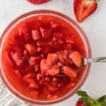 Overhead shot of a bowl of freshly made strawberry sauce with a spoon on it. Surrounding the bowl are several strawberries