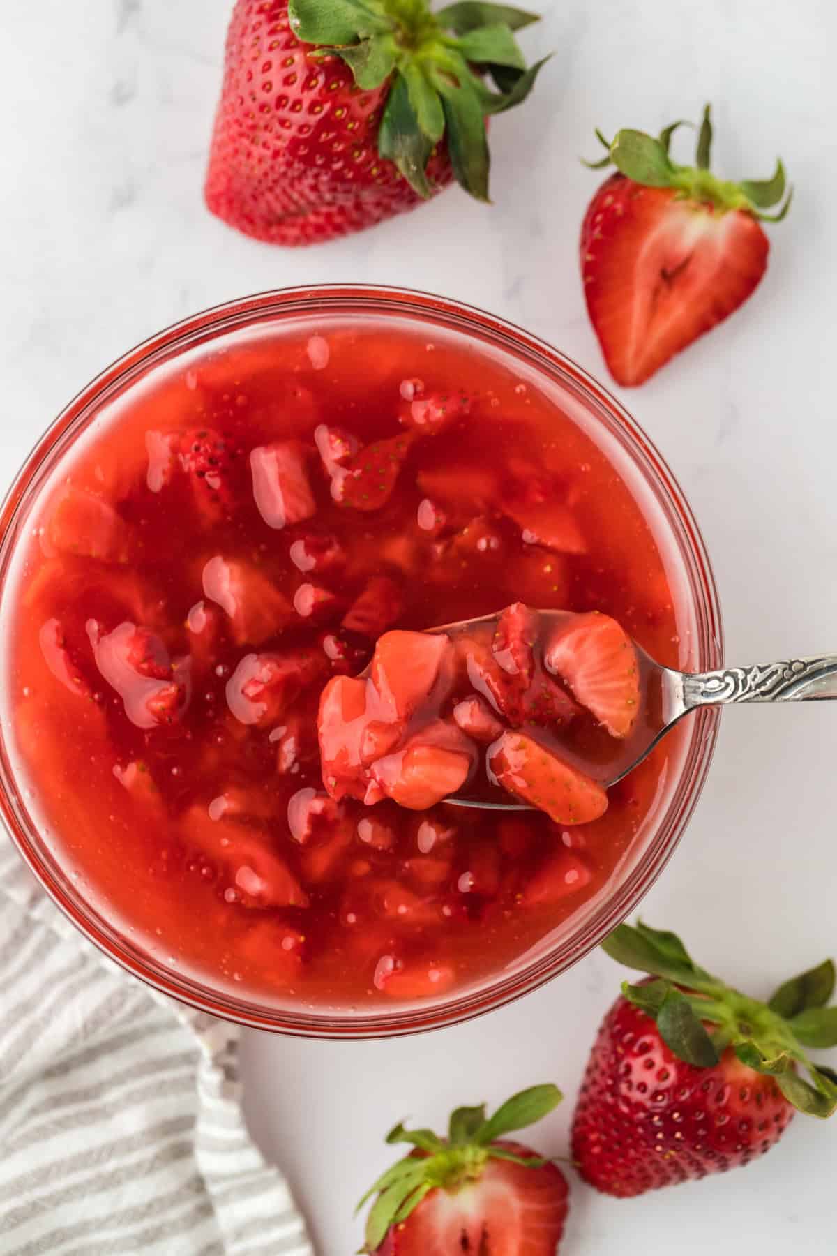 Overhead shot of a bowl of freshly made strawberry sauce with a spoon on it. Surrounding the bowl are several strawberries