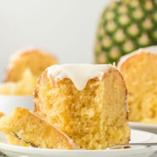 A slice of pineapple pound cake on a white plate with a fork, showing its moist interior and glaze, with a whole pineapple in the background