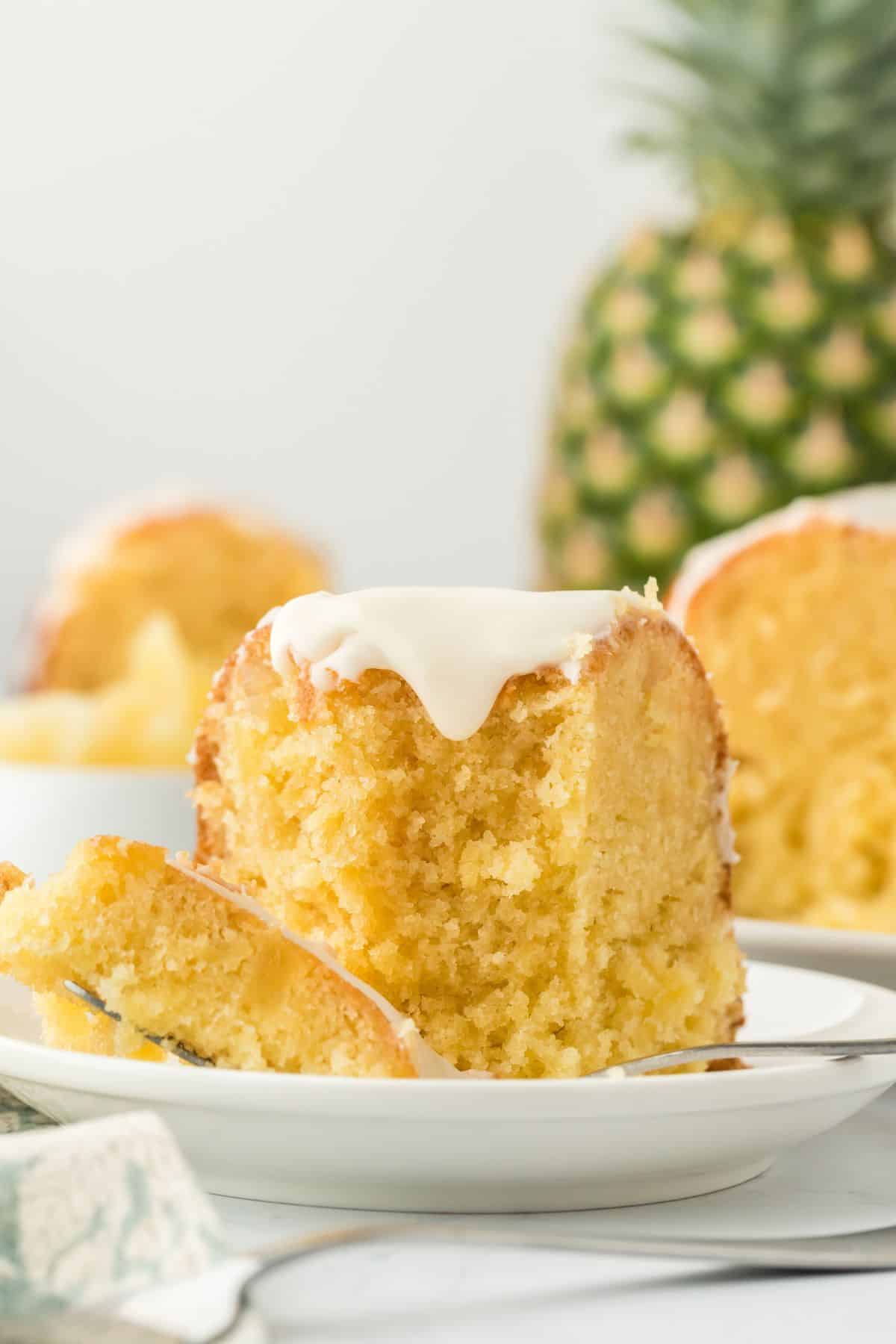 A slice of pineapple pound cake on a white plate with a fork, showing its moist interior and glaze, with a whole pineapple and more cake in the background
