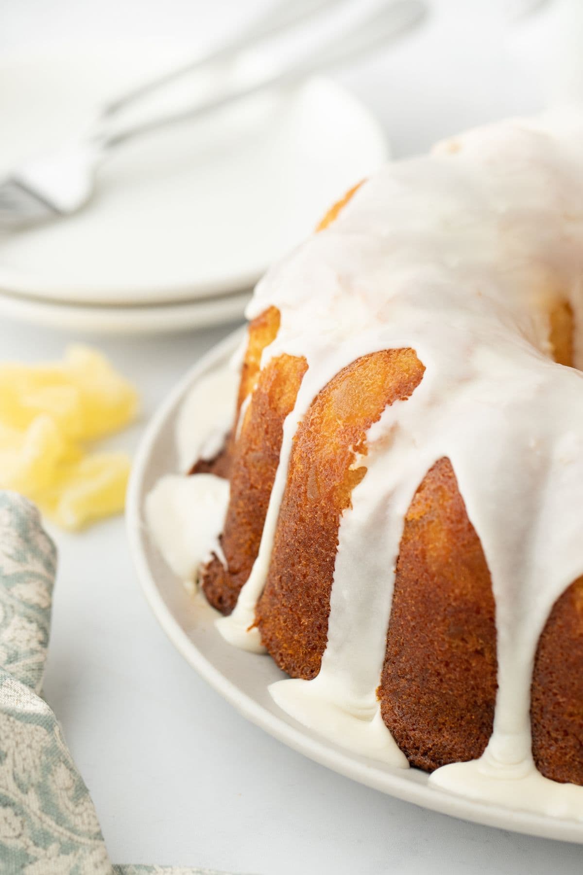 Close up of a pineapple pound cake with white glaze drizzled over it, set on a white plate with a fork and plate in the background